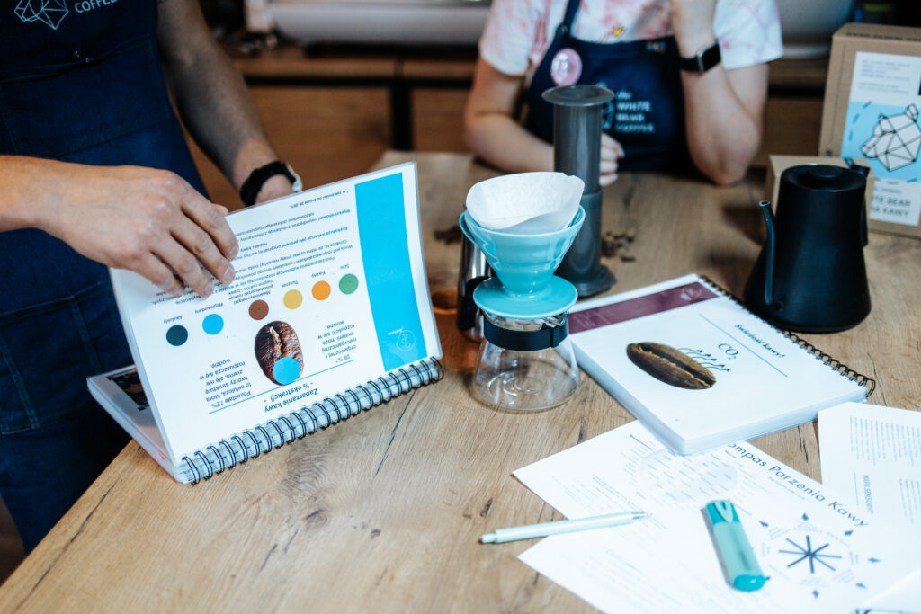 Kawa specialty. Na zdjęciu widzimy dwóch baristów przy drewnianym stole, którzy omawiają różne aspekty parzenia kawy specialty. Na pierwszym planie znajduje się niebieski dripper z papierowym filtrem oraz szklany pojemnik na zaparzoną kawę. Wokół leżą rozmaite materiały szkoleniowe, w tym otwarte książki i notatki z kolorowymi diagramami i zdjęciami ziaren kawy. Tło zdjęcia stanowi wnętrze kawiarni, co podkreśla profesjonalny charakter zajęć.