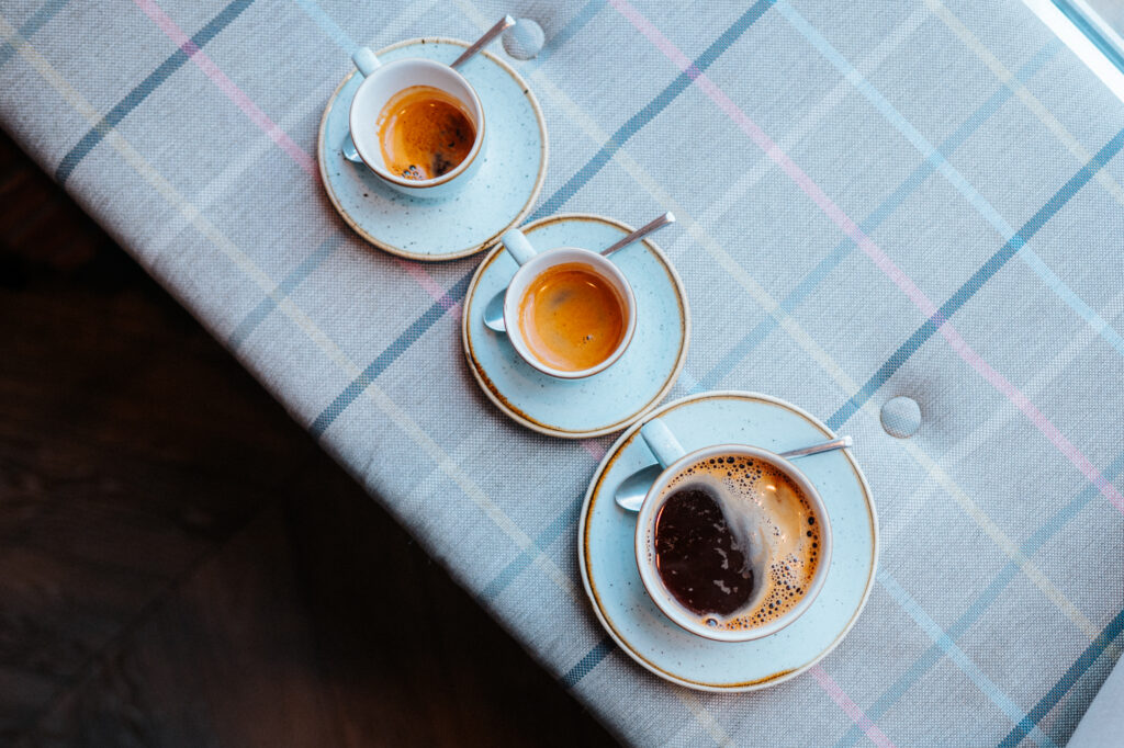 Idealne espresso. Trzy filiżanki espresso na jasnoniebieskich spodkach, ustawione na kraciastej serwetce. Każda filiżanka zawiera różne ilości kawy, od mocnego espresso po delikatniejszy napar, podkreślając różnorodność metod parzenia i intensywności espresso